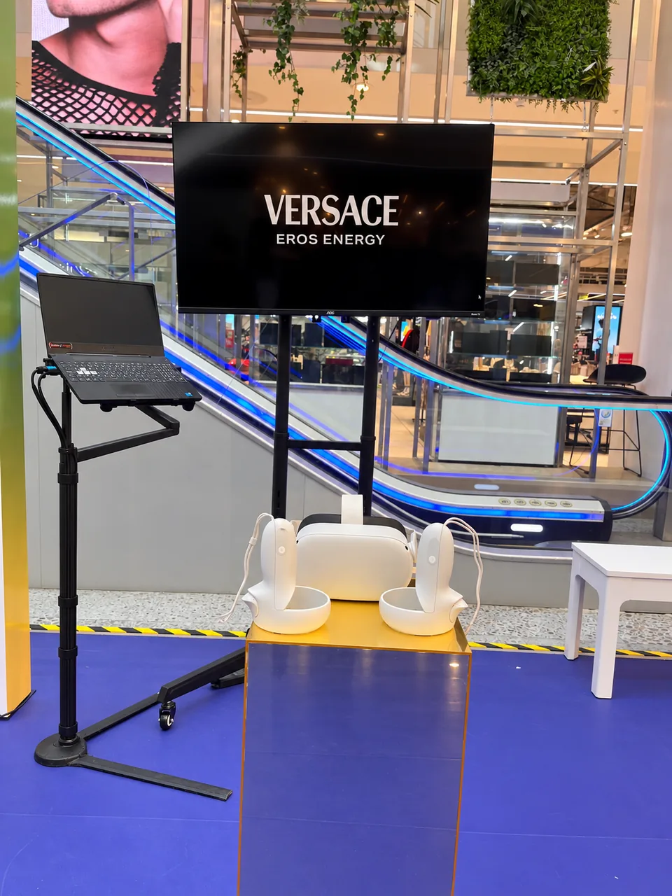 Visor y controles de realidad virtual sobre plinto espejado, notebook y pantalla de apoyo de Versace